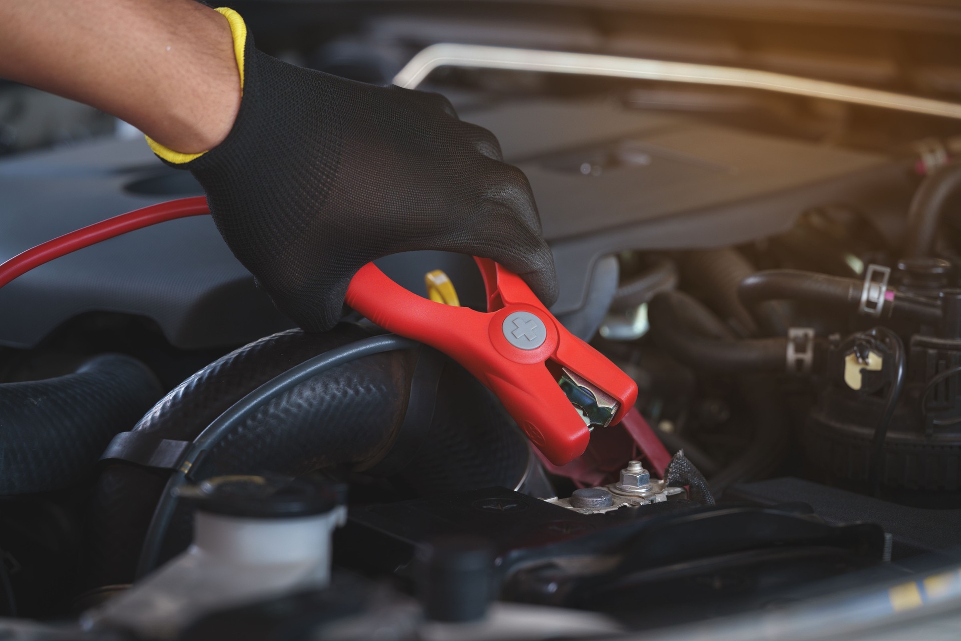 Hands in black gloves are carefully attaching red jumper cables to a car battery terminal, ready to jump-start a dead battery.
